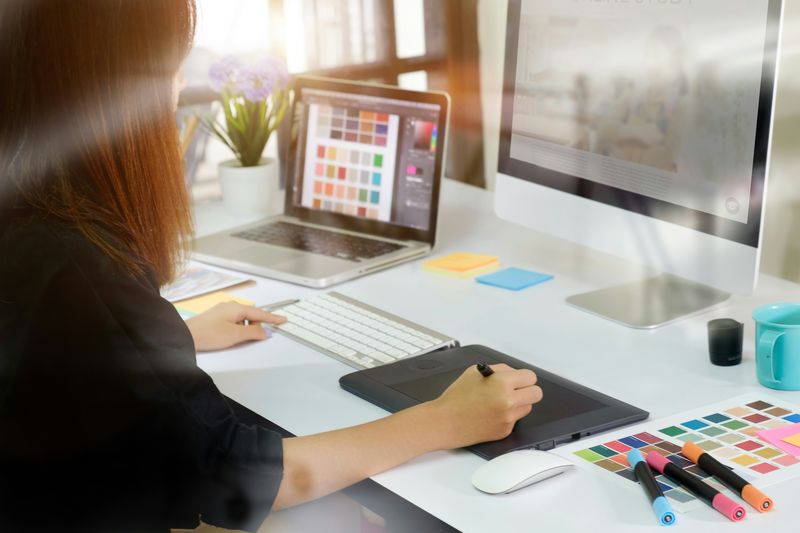 A designer working on a drawing tablet beside a laptop and colour swatches